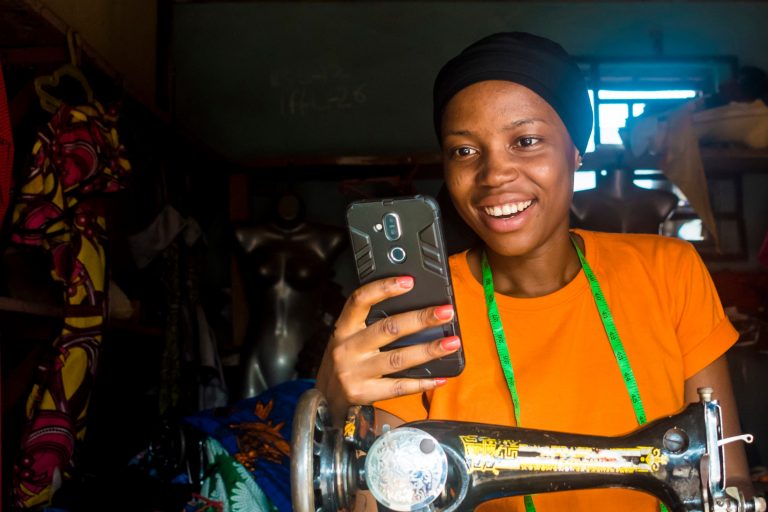 Young african female tailor smiling while viewing content on her mobile phone sitting with her sewing machine